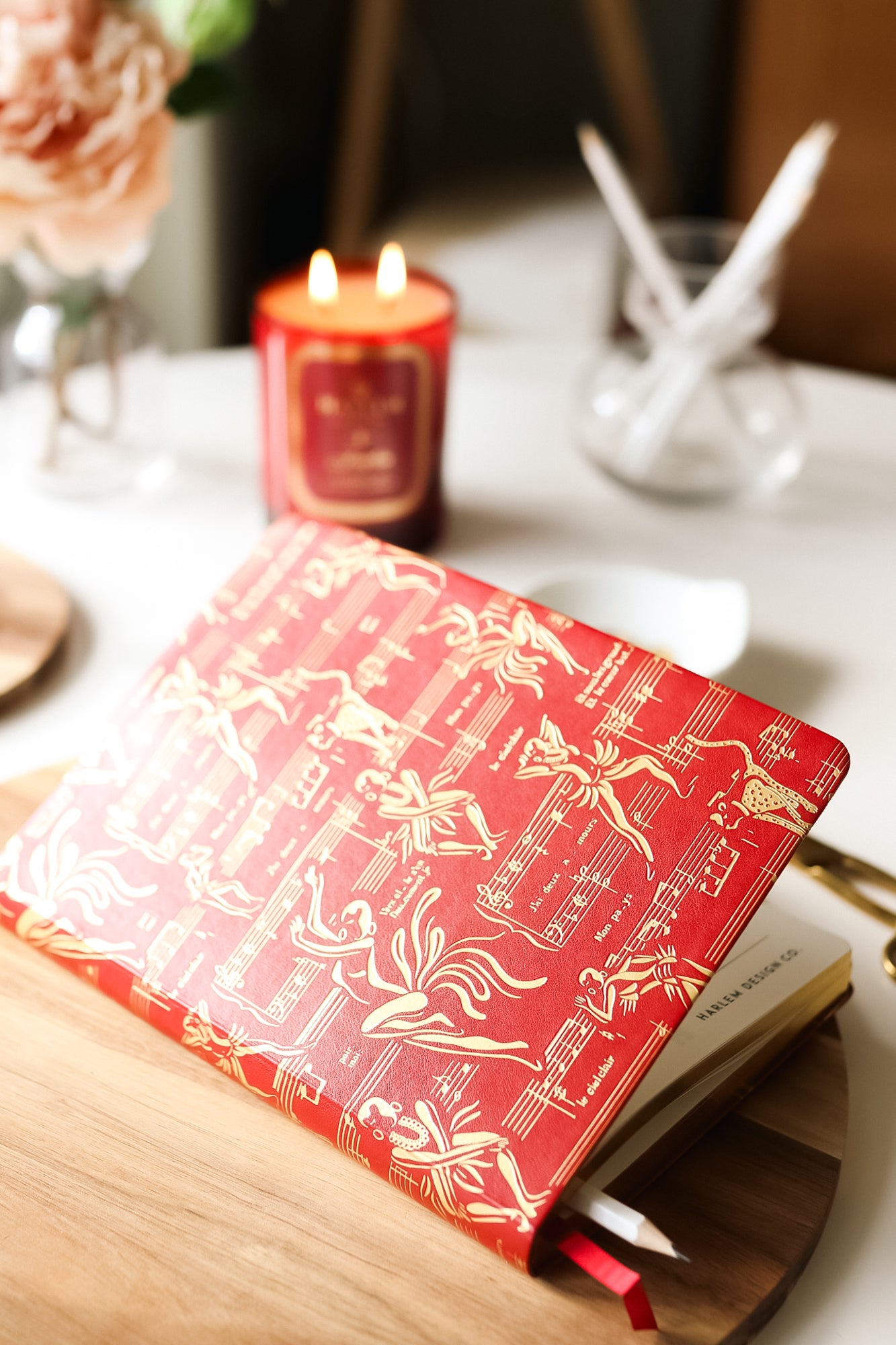 Lifestyle picture of the red Josephine journal on a wood table with the Josephine candle burning in the background. 